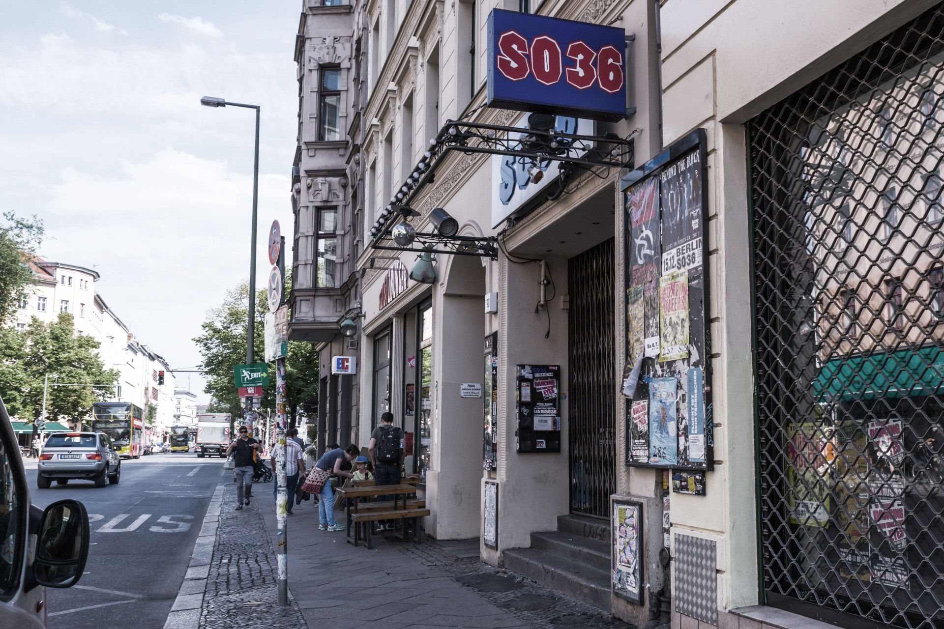 A city street scene with people by a building and a prominent sign with the number and letters "SO36" hanging above.