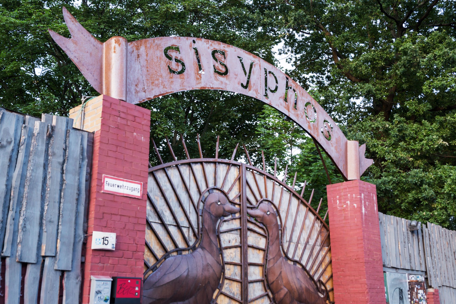 A rusted metal archway reads 'Sisyphos' flanked by sculptures of two ducks on a fence.