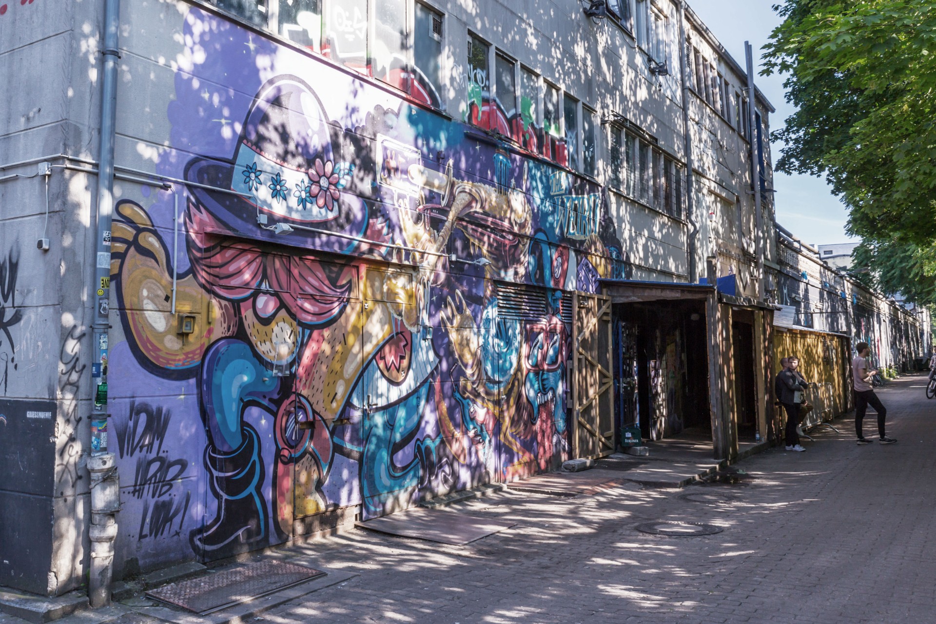 A colorful mural covers a building's facade on a sunny street with people walking by.