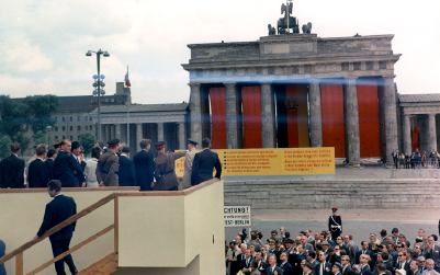A crowd gathers in front of a large building adorned with red banners and a statue. Several people stand on a makeshift platform.