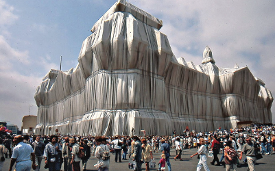 A large building is covered with white fabric, surrounded by people walking and looking.