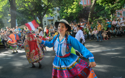 A woman in a colorful traditional outfit dances at a street parade, holding a flag.