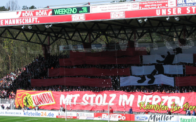 A crowd at a soccer stadium displays a large banner with text and graphic elements.