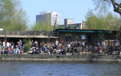 People are gathered outside a riverside café on a sunny day, enjoying the weather. Trees and modern buildings are visible in the background.