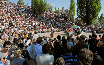 A large crowd watches a performance outdoors on a sunny day. Two performers entertain in the center of an amphitheater.