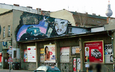 A city building facade features a large mural with a vintage car and a face, above advertisements and graffiti.