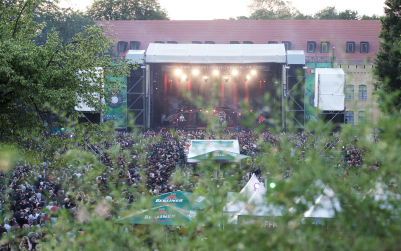 A large crowd gathers at an outdoor concert with a brightly lit stage in a park setting.