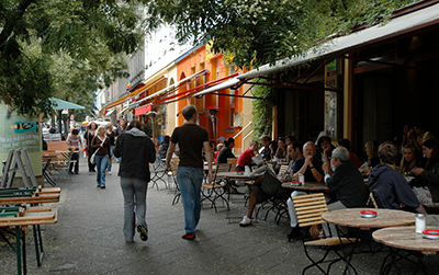 People are dining outdoors at tables along a street lined with colorful buildings.